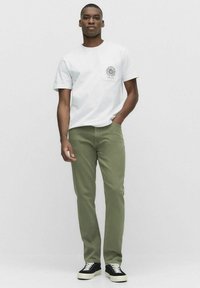 Vitt t-shirt med en bröstficka och grafisk detalj, tillsammans med gröna byxor och svarta sneakers med vita sulor. En enkel, avslappnad stil.