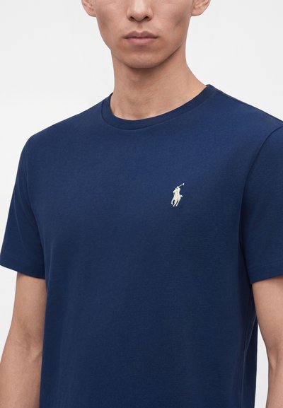 Άνδρας που φοράει μονόχρωμο σκούρο μπλε T-shirt με στρογγυλή λαιμόκοψη και μικρό λευκό λογότυπο ποδοσφαιριστή στην αριστερή πλευρά του στήθους.