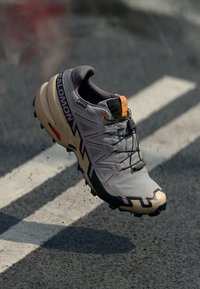 Vyriški bėgimo batai pilkos ir smėlinės spalvos su tvirtu padu, juodais akcentais ir oranžiniu ženklinimu. Lengvas medžiaga su tekstūruotais detalėmis.