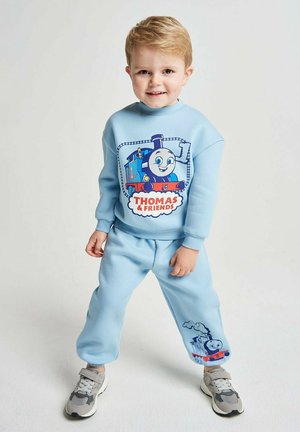 Young boy wearing light blue Thomas & Friends sweatshirt and sweatpants with sneakers, standing against plain background smiling.
