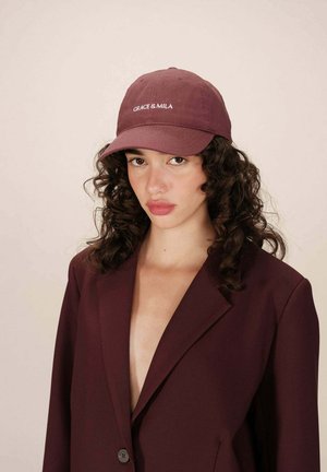 Casquette de baseball bordeaux avec "GRACE & MILA" brodé en fil blanc. Tissu lisse et visière incurvée ; le modèle porte une veste à col en V profond.