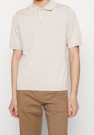 Personne portant un polo beige clair à manches courtes et un pantalon marron clair, debout devant un fond uni.