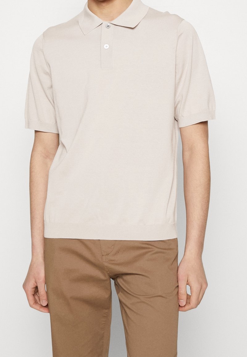 Personne portant un polo beige clair à manches courtes et un pantalon marron clair, debout devant un fond uni.