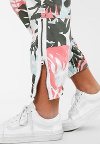 Bäddade byxor i ett flerfärgat palmmönster av grönt, rosa och blått, med dragkedjor vid anklarna och elastiska mudd. Aderade med vita sneakers.