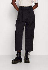 Svarta plisserade byxor med hög midja, lös passform och kort längd, kombinerade med svarta snörstövlar i kämparstil. Mjuk textur.