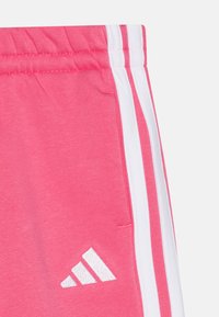 Rosa sweatshirtstof med hvid tre-stribet detalje langs siden; har et lille hvidt Adidas-logo. Inkluderer elastisk talje.