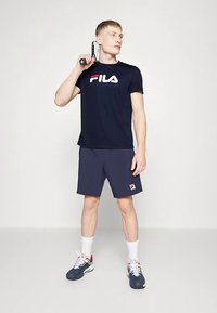 Navyblå FILA t-skjorte med logo, kombinert med mørke shorts og sportssko, holder en tennisracket, stående mot en lys bakgrunn.