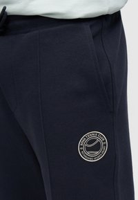 Οι βαμβακερές φόρμες σε ναυτικό μπλε διαθέτουν ανάγλυφη υφή με διαγώνιες γραμμές, πλαϊνές τσέπες και στρογγυλή λωρίδα με λογότυπο με θέμα το τένις στο μηρό.