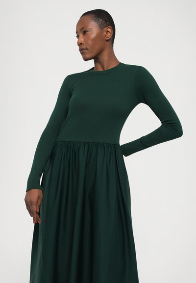 Dark green dress with a fitted long-sleeve top and a flared skirt. Smooth fabric with a subtle sheen, featuring gathered detailing at the waist.