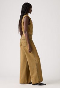 Khaki denim jumpsuit met een wijde pijpen ontwerp, mouwloze top, voorzakken en een label aan de achterkant. Soepele textuur, moderne snit en neutrale kleur.