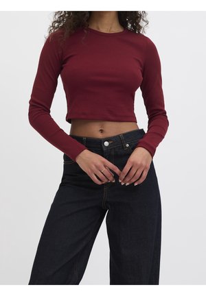Femme portant un crop top ajusté bordeaux à manches longues et un jean denim foncé taille haute, mains placées près de la ceinture.