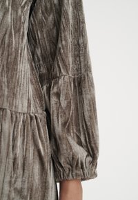 Mjukt sammets långärmad blus med samlade sömmar och elastiska ärmslut, bärs på armen mot en enfärgad vit bakgrund.