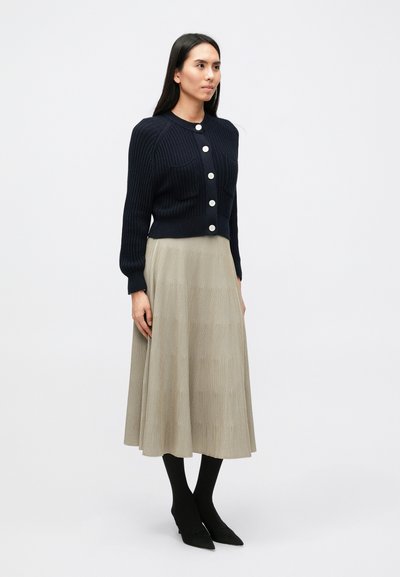 Μπλε ναυτικό ριμπ πλεκτό ζακέτα με στρογγυλή λαιμόκοψη και πέντε μεγάλα κουμπιά, συνδυασμένη με μπεζ πλισέ φούστα έως το γόνατο και μαύρα μυτερά παπούτσια.