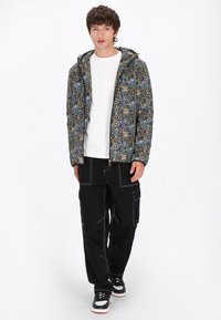 Giacca a fantasia colorata con zip, pantaloni cargo neri con cuciture bianche e scarpe nere con accenti bianchi e rossi, indossati sopra una camicia bianca.