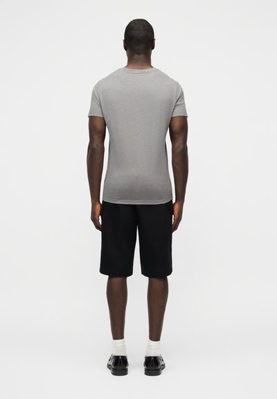 Άνδρας που στέκεται με την πλάτη προς τα εμπρός, φοράει εφαρμοστή γκρι μπλούζα, μαύρα σορτς μήκους μέχρι το γόνατο, λευκές κάλτσες και μαύρα παπούτσια.