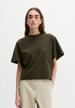 Femme aux longs cheveux bruns portant un haut ample à manches courtes vert foncé et un pantalon taille haute beige clair, debout devant un fond uni.