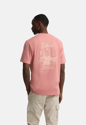 Hombre con camiseta rosa Silbon con dibujo blanco y texto "Golfe de Saint-Tropez" en la espalda, combinado con pantalones cargo beige, de espaldas.