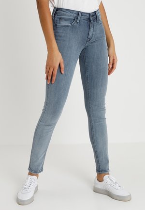 Kobieta w jasnoniebieskich obcisłych dżinsach i białych trampkach, stojąca ze swobodnie opuszczonymi rękami wzdłuż ciała na jednolitym tle.