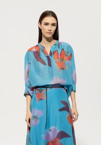 Blaues Blumenbluse mit lockerer Passform, die kräftige orange und lila Muster aufweist, mit einer Knopfleiste vorne und gerafften Ärmeln, kombiniert mit einem passenden Rock.