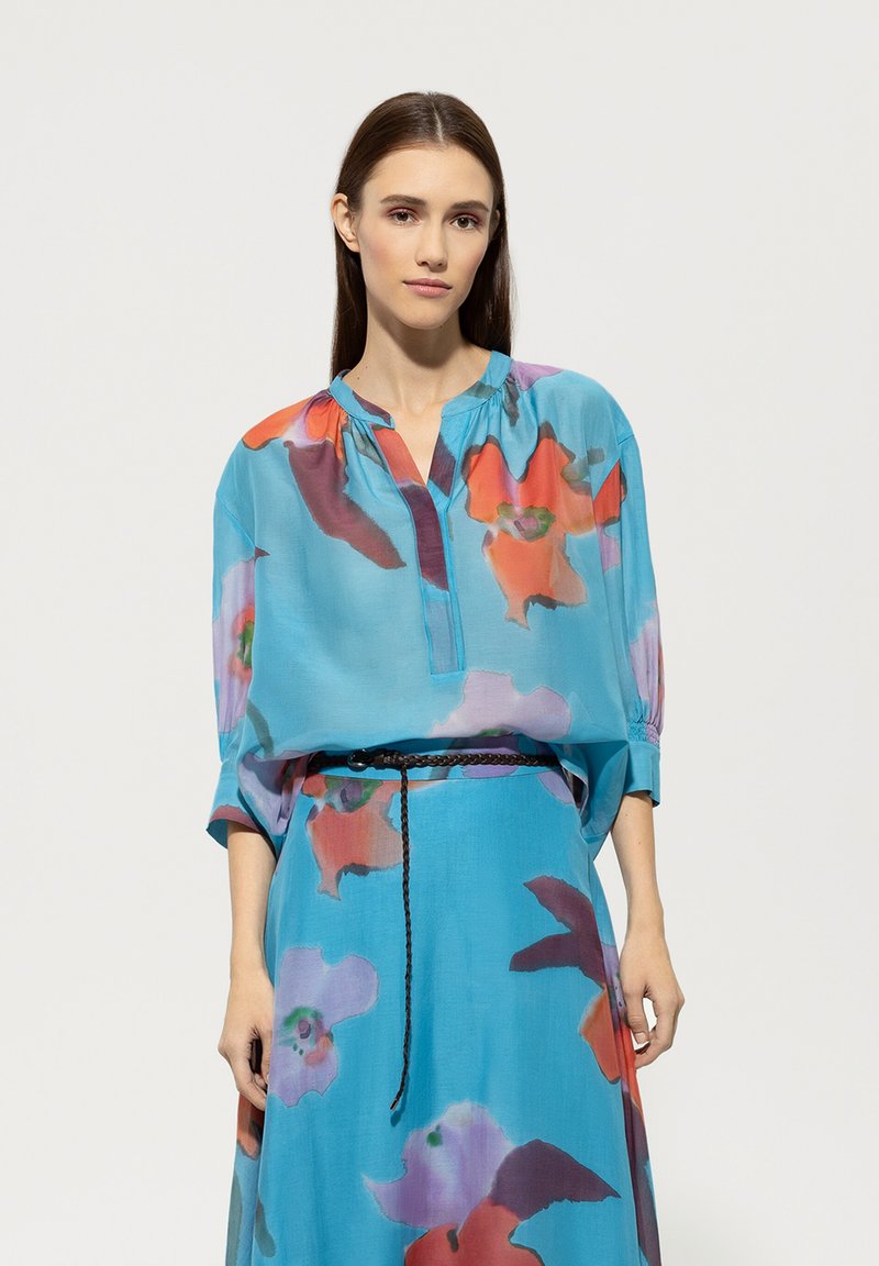 Blaues Blumenbluse mit lockerer Passform, die kräftige orange und lila Muster aufweist, mit einer Knopfleiste vorne und gerafften Ärmeln, kombiniert mit einem passenden Rock.