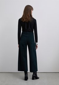 Haut noir à manches longues avec un nœud au dos, associé à un pantalon large en velours côtelé vert foncé et des bottines noires à talons carrés.