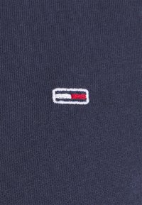 Marinblått bomullstyg med en liten broderad logotyp i röd, vit och blå färg, placerad nära det nedre vänstra hörnet.