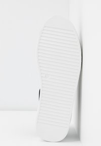Semelle d'une basket blanche en caoutchouc avec des rainures horizontales, reposant verticalement contre un mur blanc.