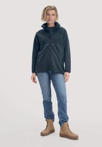 Donna in piedi che indossa un fleece grigio scuro con zip, jeans blu e stivali con plateau beige contro uno sfondo chiaro e uniforme.