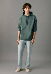 Sudadera verde con capucha, bolsillo canguro y cordones, combinada con jeans azul claro y zapatillas beige. Diseño simple, ajuste informal.