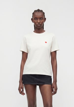Mujer con el cabello trenzado, vistiendo una camiseta blanca con un pequeño corazón rojo en el pecho y una minifalda de mezclilla negra, de pie frente a un fondo liso.