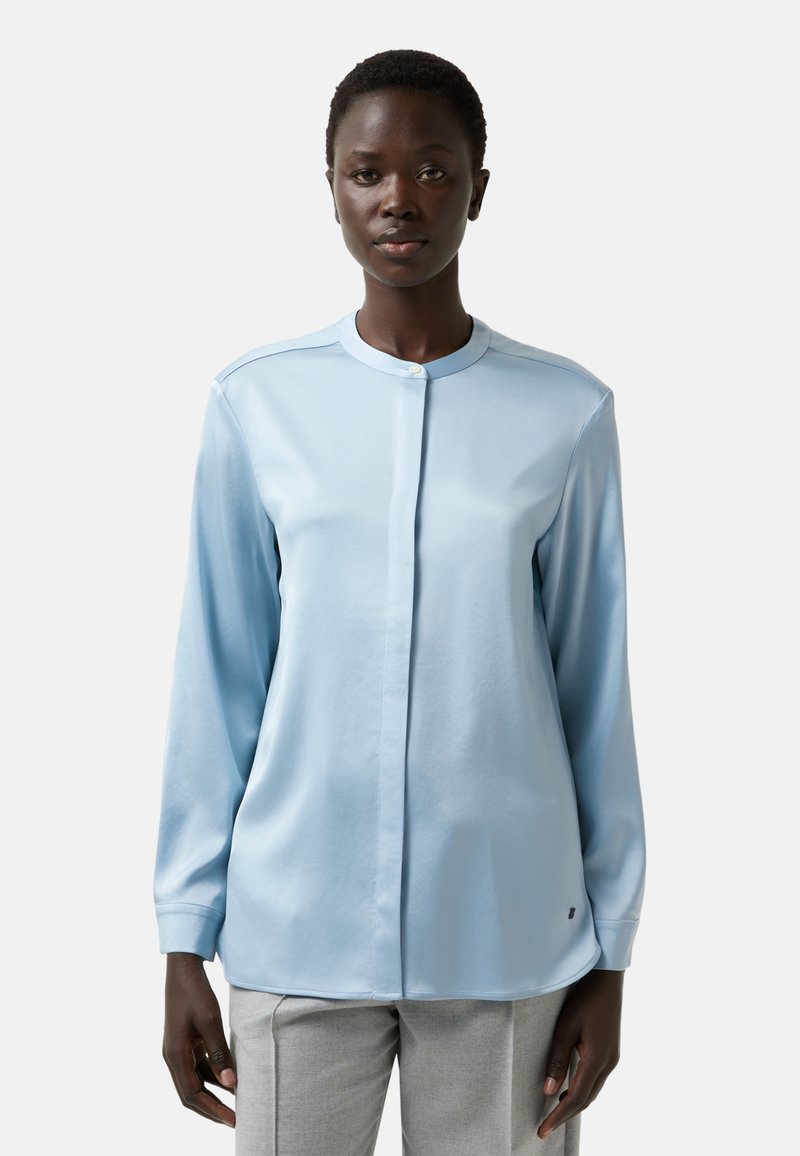 Blouse bleu clair à manches longues avec un col rond et fermeture à boutons. Tissu lisse avec un léger éclat, associé à un pantalon gris clair.
