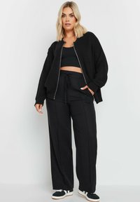 Svart oversized ribbad jacka med dragkedja, tillsammans med en åtsittande svart crop top och matchande vida byxor. Svarta sneakers fulländar looken.