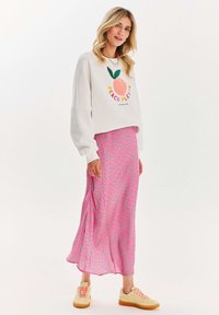 Woman wearing white sweatshirt with peach graphic, pink and green spotted long skirt, and cream sneakers with pink laces, standing on white background.