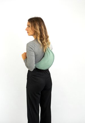 Sac en tissu vert porté sur le dos, léger et à la forme courbée. Le modèle porte un haut gris à manches longues et un pantalon noir. Style simple et fonctionnel.