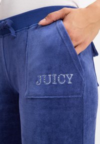Purple velour joggers feature a drawstring waistband and two pockets, with "JUICY" embroidered in crystals on the left pocket.