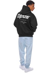 Svart hettegenser med hvit tekst på ryggen, som viser "Dpszé" og "Elite League 2020." Kombinert med lyseblå jeans og hvite sneakers.