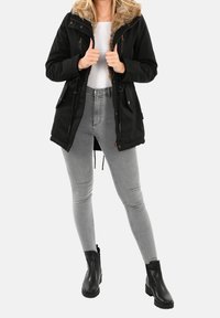 Parka noire avec une capuche en fourrure, dotée de cordons de serrage réglables, de poches zippées et d'une silhouette légèrement évasée. Portée sur un jean gris et des bottines noires.