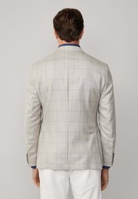 Mężczyzna odwrócony tyłem, ubrany w jasnoszary żakiet w niebieskie kraty, ciemnoniebieską koszulę i białe spodnie na jednolitym tle.