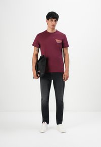 Junger Mann trägt ein kastanienrotes Tommy Jeans T-Shirt, schwarze Jeans, weiße Sneaker und hält eine schwarze Tasche vor einem weißen Hintergrund.