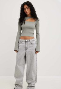 Top de manga larga acanalado en verde apagado con un corte en el pecho y mangas alargadas, combinado con jeans sueltos de corte claro.