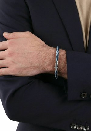 Hombre con traje oscuro y camisa beige cruza los brazos, mostrando una pulsera tejida azul y gris con cierre de plata en la muñeca.