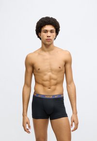 Mustad Nike bokserid elastse vöökohaga, millel on oranži logotekst. Modell on sale ja seisab ühevärvilise valge tausta ees.