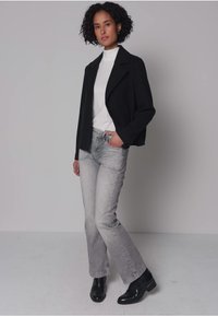 Zwarte cropped blazer over een witte coltrui, gecombineerd met lichtgrijze flare jeans en zwarte enkellaarzen. Getextureerde stof en eenvoudig ontwerp.