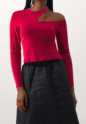 Vrouw die een rode top met lange mouwen draagt met een asymmetrische halsuitsnijding en een zwarte, doorgestikte hoge taille rok met elastische tailleband.