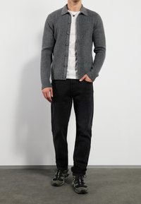 Graue gestrickte Strickjacke mit Kragen, Knopfleiste, getragen über einem weißen T-Shirt, kombiniert mit schwarzen Hosen und schwarzen Turnschuhen.