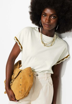 Femme avec un grand afro, portant une chemise blanche à bordure en dentelle, des colliers superposés, un pantalon crème et tenant un sac à main en daim beige sur fond blanc.