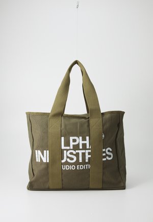 Sac fourre-tout en toile vert olive avec des poignées épaisses et un texte blanc « Alpha Industries Studio Edition » sur le devant, suspendu sur un fond uni.