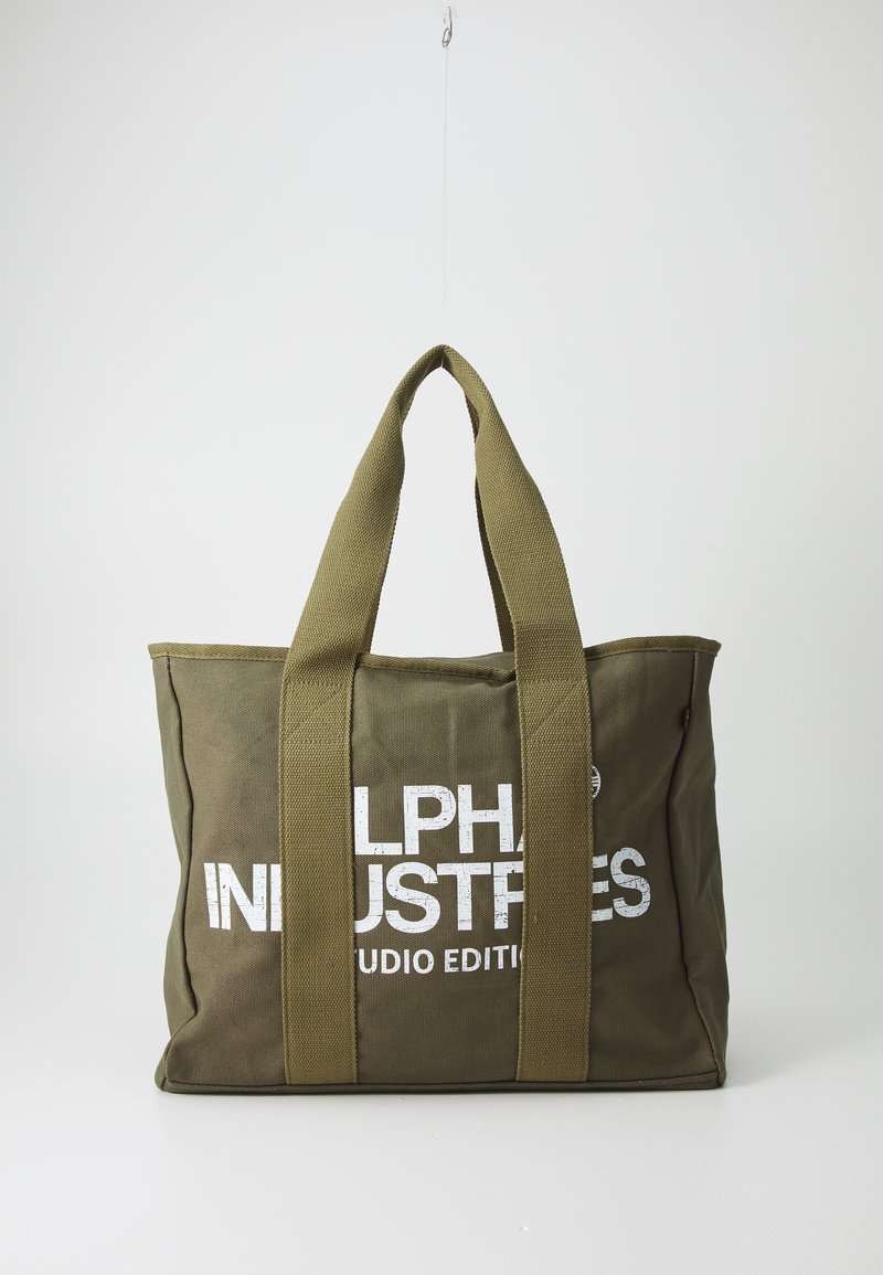 Bolsa tote de lona verde oliva con asas gruesas y texto blanco "Alpha Industries Studio Edition" en el frente, colgada contra un fondo liso.