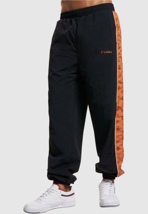 Sorte joggingbukser med orange sidebånd, der har tekst og grafik. Elastisk talje og manchetter ved anklerne. Sammen med hvide sneakers.