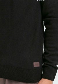 Schwarzer Strickpullover mit strukturiertem Material und gerippten Bündchen, ausgestattet mit einem kleinen braunen Etikett am unteren Saum mit dem Markennamen.